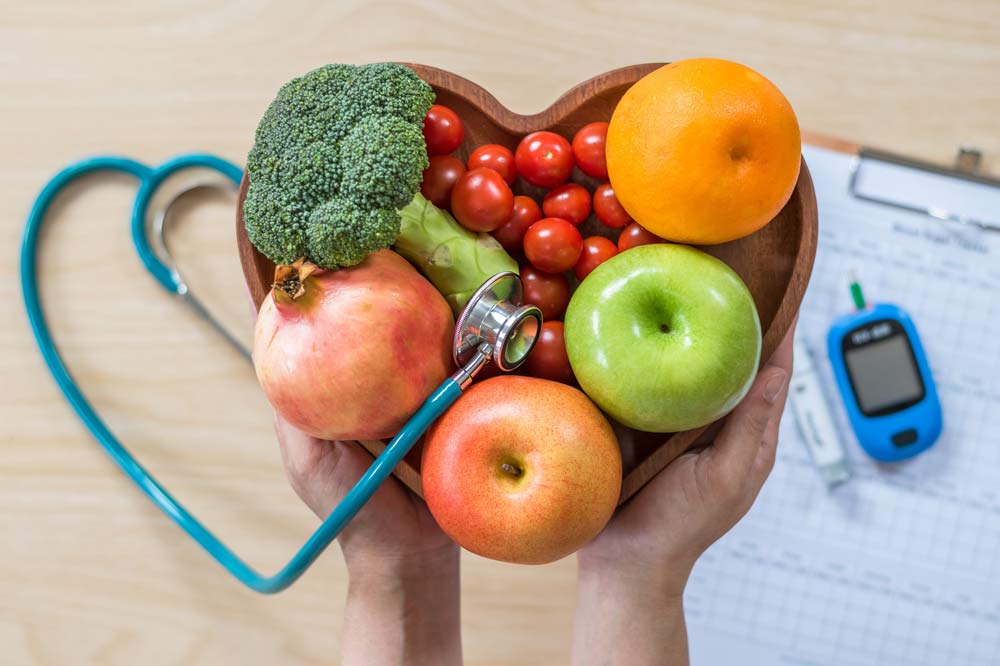 Eine Person hält eine herzförmige Schale mit Obst und Gemüse. Rechts daneben liegt ein Blutzuckermessgerät und links ein Stethoskop