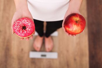 Молодая женщина стоит на весах, держа в одной руке яблоко, а в другой — пончик.
