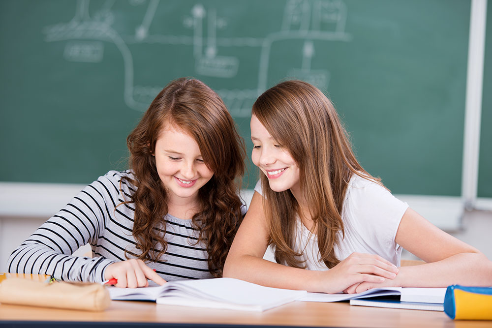 2 Kinder sitzen in einem Klassenzimmer vor einer Tafel und schauen zusammen in ein Buch.