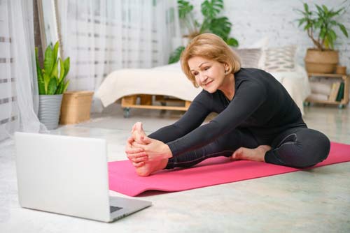 A woman doing stretching exercises in her home while looking at a laptop.