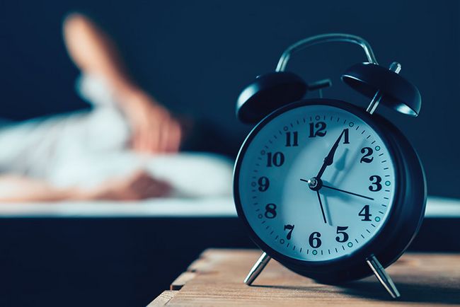 Ein Wecker zeigt 5:05 Uhr an. Im Hintergrund ist schemenhaft eine Person in einem Bett zu sehen.