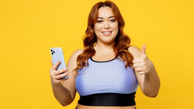 Übergewichtige Frau, die in die Kamera lächelt und ein Smartphone hält