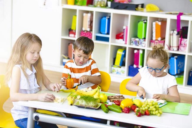 Drei Grundschulkinder sitzen an einem Tisch in einem Klassenzimmer und schneiden Obst und Gemüse.