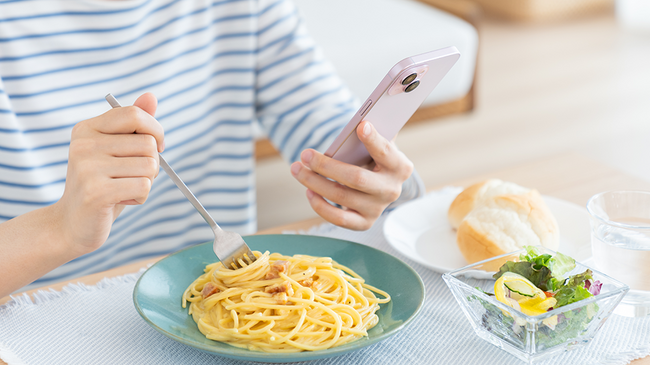 Eine Frau sitzt am Tisch, hat einen Teller Spaghetti vor sich und tut sich mit der einen Hand eine Gabel davon auf, während sie ein Smartphone in der anderen Hand hält.
