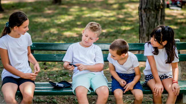 Ein Junge sitzt mit anderen Kindern auf einer Parkbank und erklärt ihnen seine Erkrankung