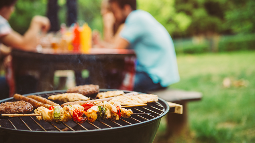 Im Vordergrund ist ein Grillrost mit Gemüsespießen und Fleisch zu sehen, im Hintergrund sitzen Menschen an einem Tisch im Park. Auf dem Tisch stehen Saucen.