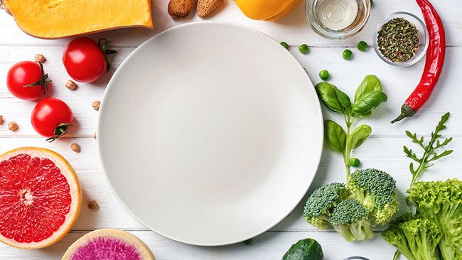 Ein leerer Teller steht in der Mitte des Bildes. Um den Teller herum liegen gesunde pflanzenbasierte Lebensmittel.
