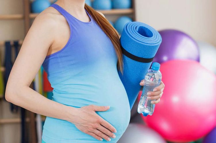 Eine schwangere Frau steht in einem Sportraum mit einer Yogamatte unter dem Arm.