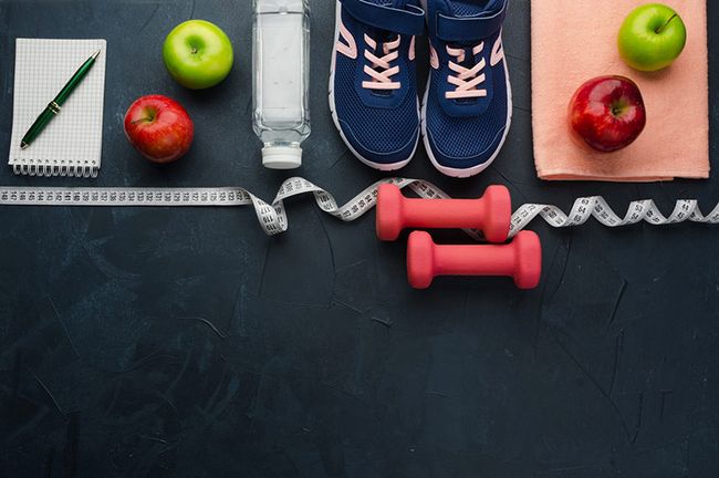 In einer Reihe liegen ein Block mit Stift, Äpfel, eine Wasserflasche, Sportschuhe, Hanteln und ein Maßband.