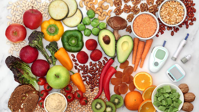 Auf einem Tisch liegen verschiedene Hülsenfrüchte, Nüsse, Obst- und Gemüsesorten zusammen mit einem Glukosemessgerät und einem Schrittzähler.