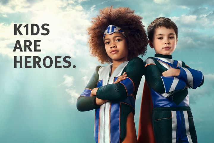 Zwei Kinder in Superheldenkostüm sind auf einer Plakatwand zu sehen.