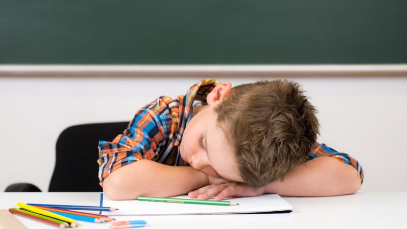 Ein junge sitzt müde an einem Tisch in der Schule und hat den Kopf auf die Arme gelegt. 