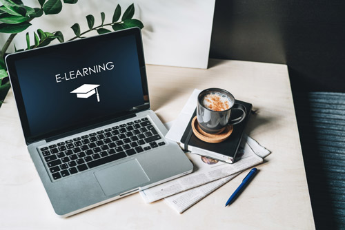 Auf einem Laptop steht "E-Learning". Neben dem Laptop steht eine Tasse Kakao auf Zeitungen.